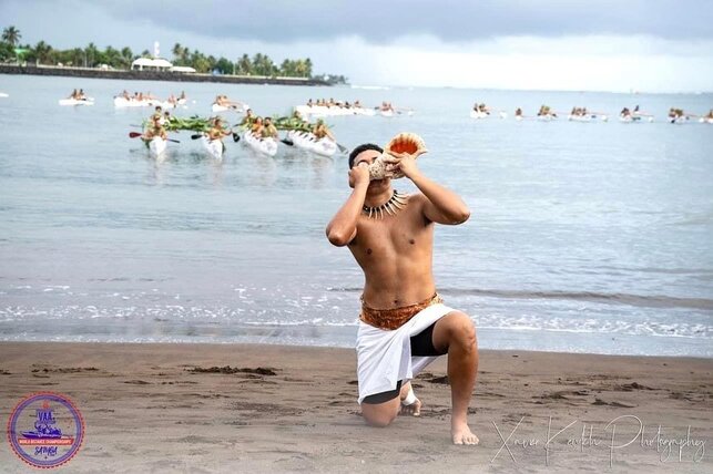 A Samoan man half kneels on a beach with outrigger canoes behind him. He wears traditional dress and blows a conch shell.