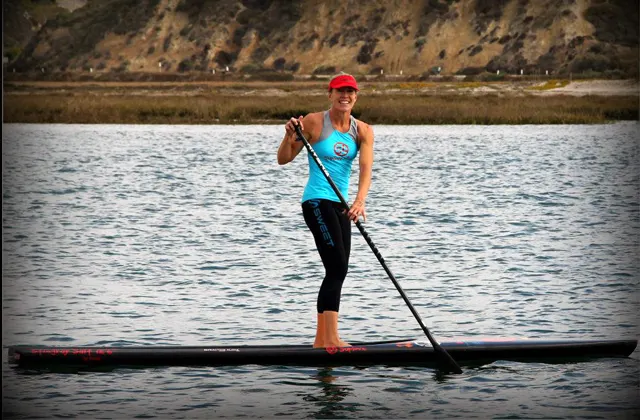 Kristin Thomas standing on a paddleboard, doing a cross-bow stroke while turning to face the camera smiling